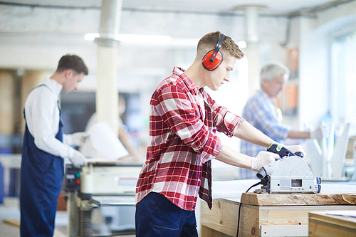 Serious concentrated young male carpenter in ear protectors wearing checkered shirt and work gloves standing at desk and working with circular saw while cutting wooden plank in workshop