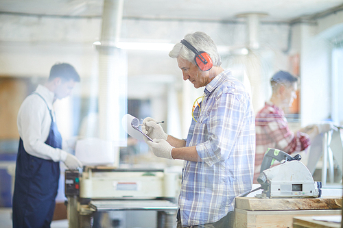 Serious thoughtful handsome mature carpenter in ear protectors leaning on workbench and analyzing notes in clipboard while standing in workshop