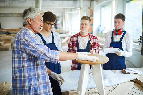 Serious confident senior carpenter in gloves touching wooden stool while teaching young students to make furniture from wood at class