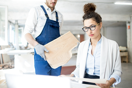 Young serious female analyst looking at data on laptop display with engineer holding wooden board on background