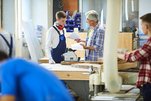 Irritated emotional senior foreman standing at workbench with polish machine and yelling at inexperienced carpenter for mistakes in sketches in factory workshop