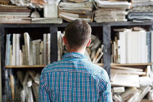 Back view of contemporary artisan standing in front of shelves with patterns or workpieces in his workshop