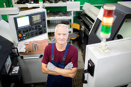 Portrait of confident mature operator standing in overall with arms crossed standing at workplace among computer machines
