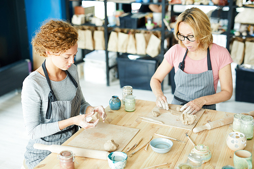 Young woman and her colleague in aprons sitting by wooden table and working with clay workpieces