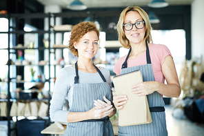 Two happy young creative females in aprons looking at you in workshop