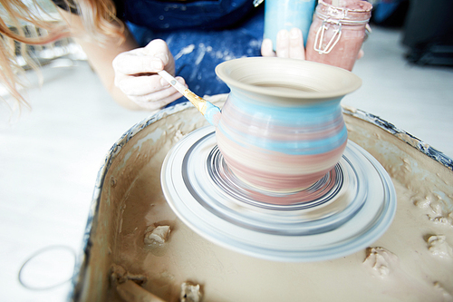 Modern potter touching rotating jug by paintbruh with blue color during process of creation