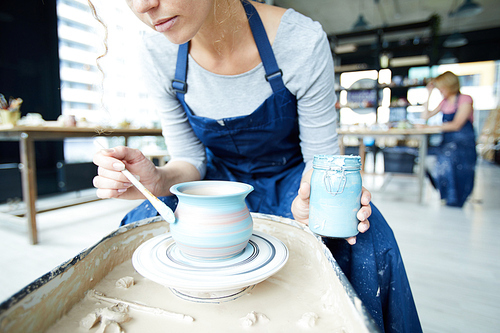 Female potter with jar of blue color sitting by rotating clay jug and painting it
