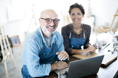 Smiling aged man and his colleague in workwear sitting by table in front of laptop in their workshop or studio