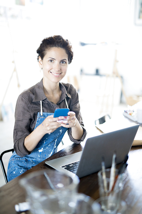 Young smiling creative woman sitting by table in studio and texting in smartphone