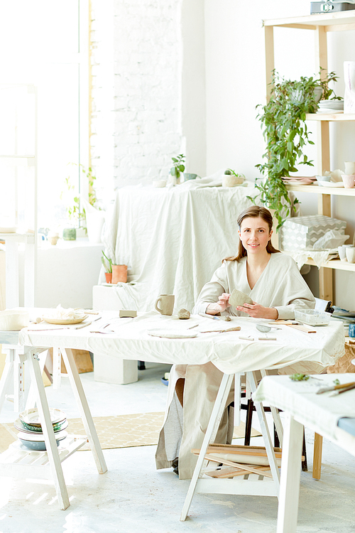 Happy young female in workwear sitting by table in her workshop,  and making kitchenware from clay