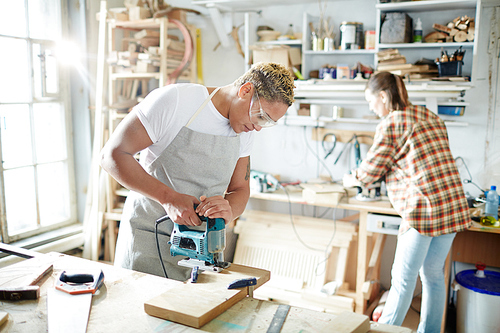 Young carpenter with electric fretsaw sawing wooden board in workshop