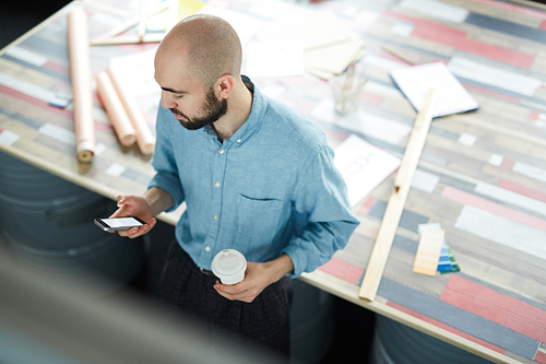Directly above view of serious handsome young bearded designer in blue shirt standing at desk and checking message on smartphone while drinking coffee at workplace