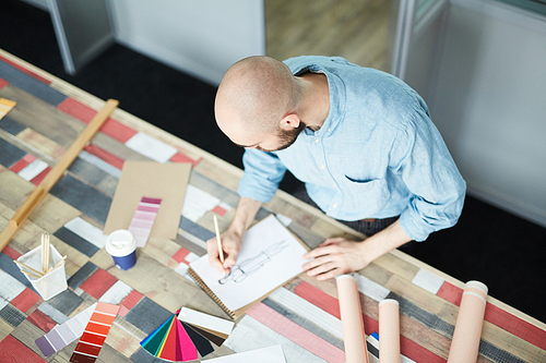 Serious male creative fashion designer in blue shirt standing at table with color swatches and papers and making new sketch of dress in studio