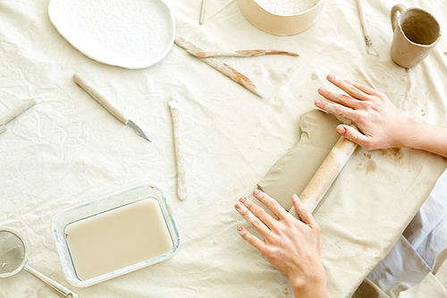 Handcraft master making piece of white clay flat with help of wooden rolling-pin on table