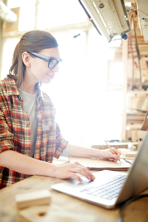 Beautiful woman in glasses using laptop to make calculations while working in carpenter workshop.