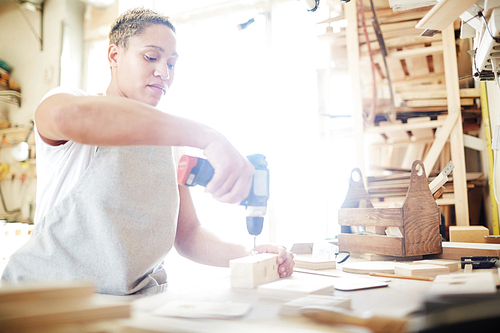 Carpenter with electric drill working with wooden plank by workbench