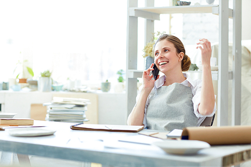 Young creative woman in workwear sitting in her workroom while speaking on smartphone and sharing good mood with friend