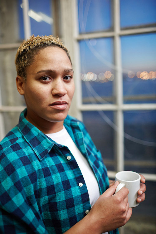 Adult gritty woman holding mug of hot beverage and  while standing near window in workshop.