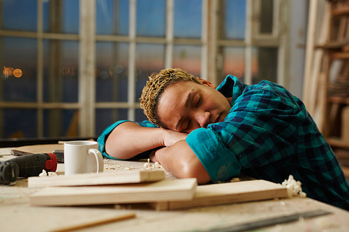 Exhausted carpenter sleeping by workplace with mug and wooden materials near by