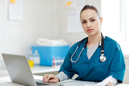 Young female veterinarian in uniform sitting by desk in front of laptop and 