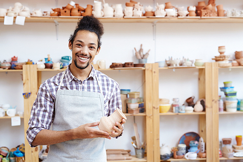 Portrait of young mixed-race man in apron holding handmade clay vase in workshop and smiling at camera cheerfully