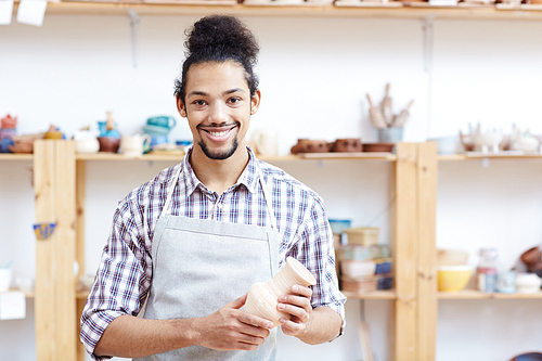Portrait of young mixed race man in apron standing in workshop with handmade clay vase and smiling at camera happily