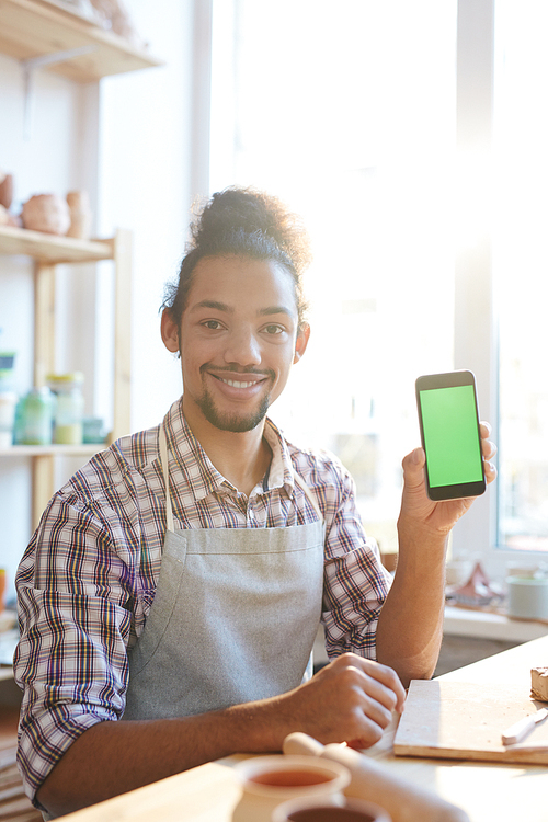 Portrait of young mixed race craftsman in apron sitting at table in workshop, holding smartphone and smiling at camera