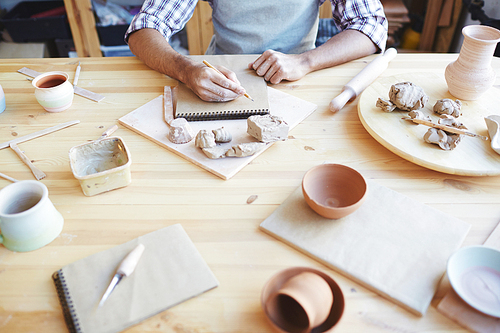 Unrecognizable male potter drawing in sketchbook while sitting at messy table with clay handicrafts in workshop