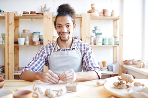 Portrait of young handsome mixed race craftsman smiling at camera while sitting at table and shaping clay in workshop