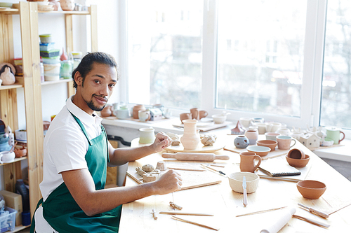 Portrait of young African American male potter sitting at messy table with handicrafts and 