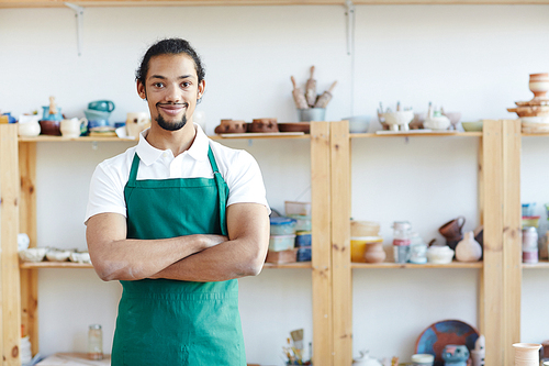 Portrait of handsome mixed race craftsman standing in pottery workshop with crossed hands and smiling at camera