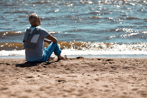 Aged relaxed man sitting on sandy beach by coastline and taking pleasure in summer day