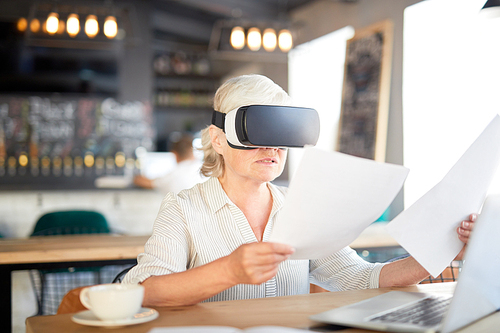 Modern senior financier with papers watching virtual presentation in vr goggles while sitting in cafe