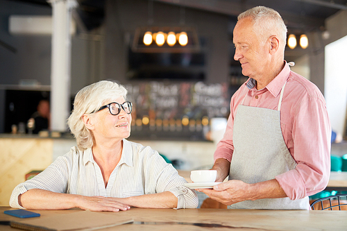 Senior waiter in apron bringing cup of tea or coffee to mature visitor in cafe or restaurant