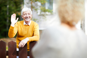 Cheerful and friendly mature man waving hand to his neighbour while standing by fence