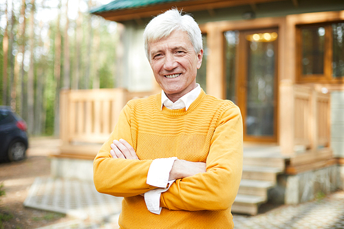 Mature man with grey hair crossing his arms on chest while standing ny his country house or cottage