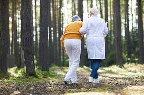 Female clinician in whitecoat helping sick man to walk while both moving down forest path