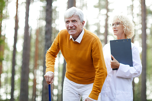 Mature man with sick leg limping while moving in natural environment with clinician near by