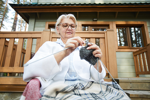 Mature woman relaxing in rocking chair by terrace of cottage on summer day and knitting socks for grandchildren