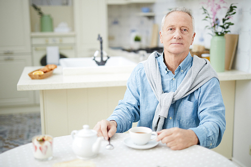 Mature man in casualwear sitting by table in the kitchen and having tea in the morning