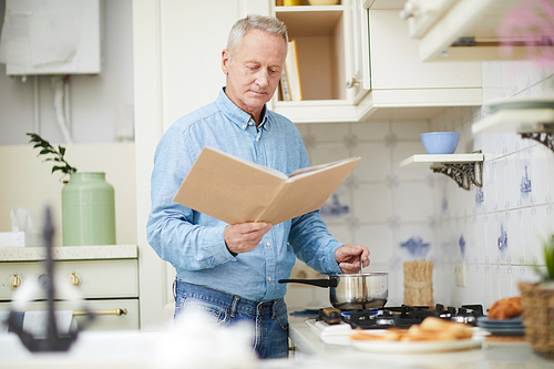 Mature man with open cookery book standing by stove and mixing something in pan