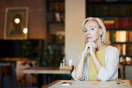 Mature blonde woman in smart casual sitting by table in cafe and looking at you