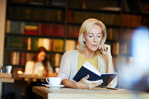 Pensive blonde mature woman sitting by desk in library or cafe and reading book by cup of tea