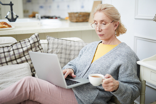 Restful aged woman with laptop and cup of tea sitting on sofa, networking or watching webcast
