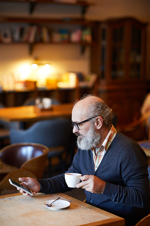 Senior bearded man with gadget and cup of tea or coffee sitting by table and texting in cozy cafe