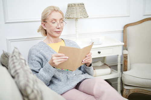Contemporary aged female with open book sitting on sofa in living-room and reading at leisure