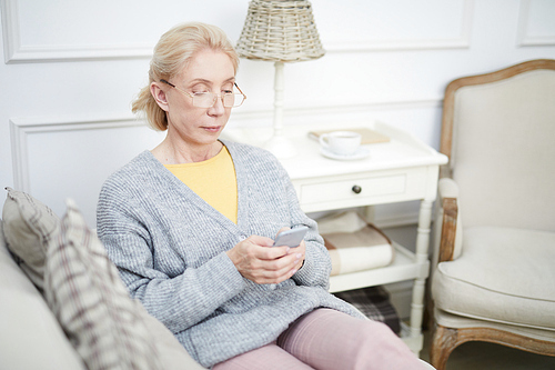 Senior female in domestic clothes sitting on sofa in living-room and reading messaging in her smartphone