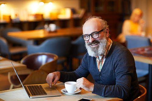 Cheerful senior man in casualwear looking at you while sitting by table in front of laptop and having tea