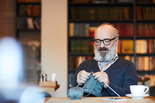 Bearded retired man in casualwear and eyeglasses knitting warm woolen winterwear