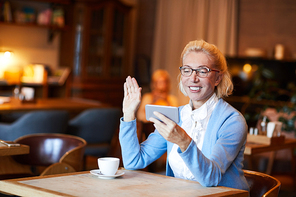 Cheerful blonde mature female waving hand to someone in her smartphone while talking through video chat